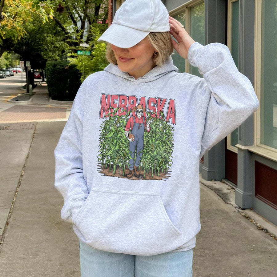 Nebraska With A Skeleton Dressed As A Farmer In Overalls Holding A Football And An Ear Of Corn While Standing In A Cornfield Hoodie