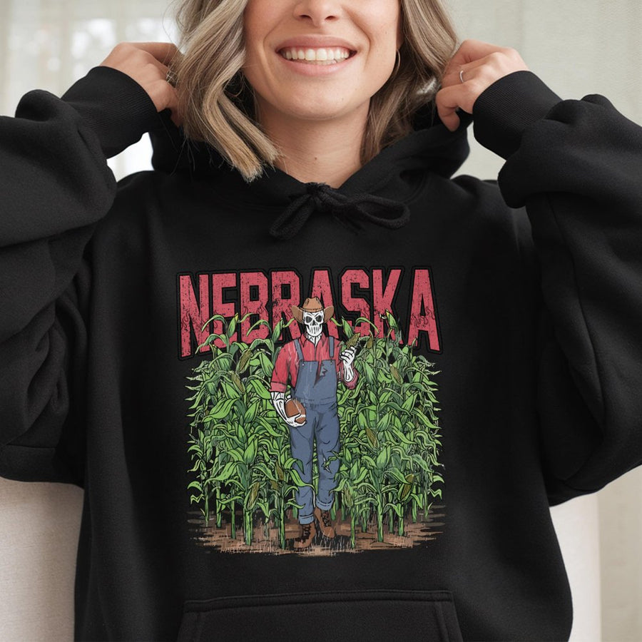 Nebraska With A Skeleton Dressed As A Farmer In Overalls Holding A Football And An Ear Of Corn While Standing In A Cornfield Hoodie