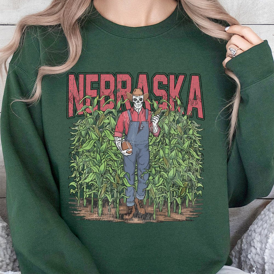 Nebraska With A Skeleton Dressed As A Farmer In Overalls Holding A Football And An Ear Of Corn While Standing In A Cornfield Sweatshirt