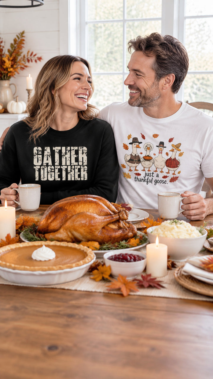 Couple laughing at a Thanksgiving dinner table, woman in a black Gather Together sweatshirt, man in a white thankful goose t-shirt