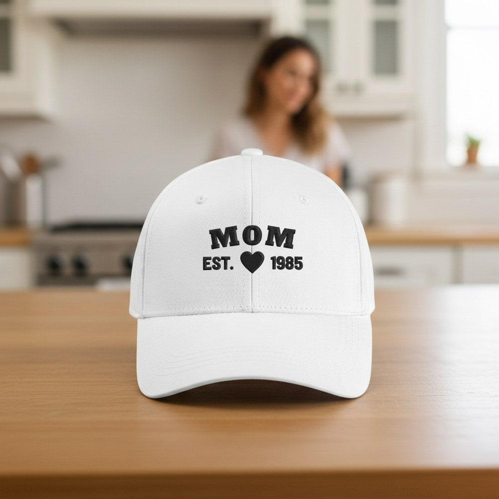White cap embroidered MOM EST. 1985 with a heart, on a bright kitchen table